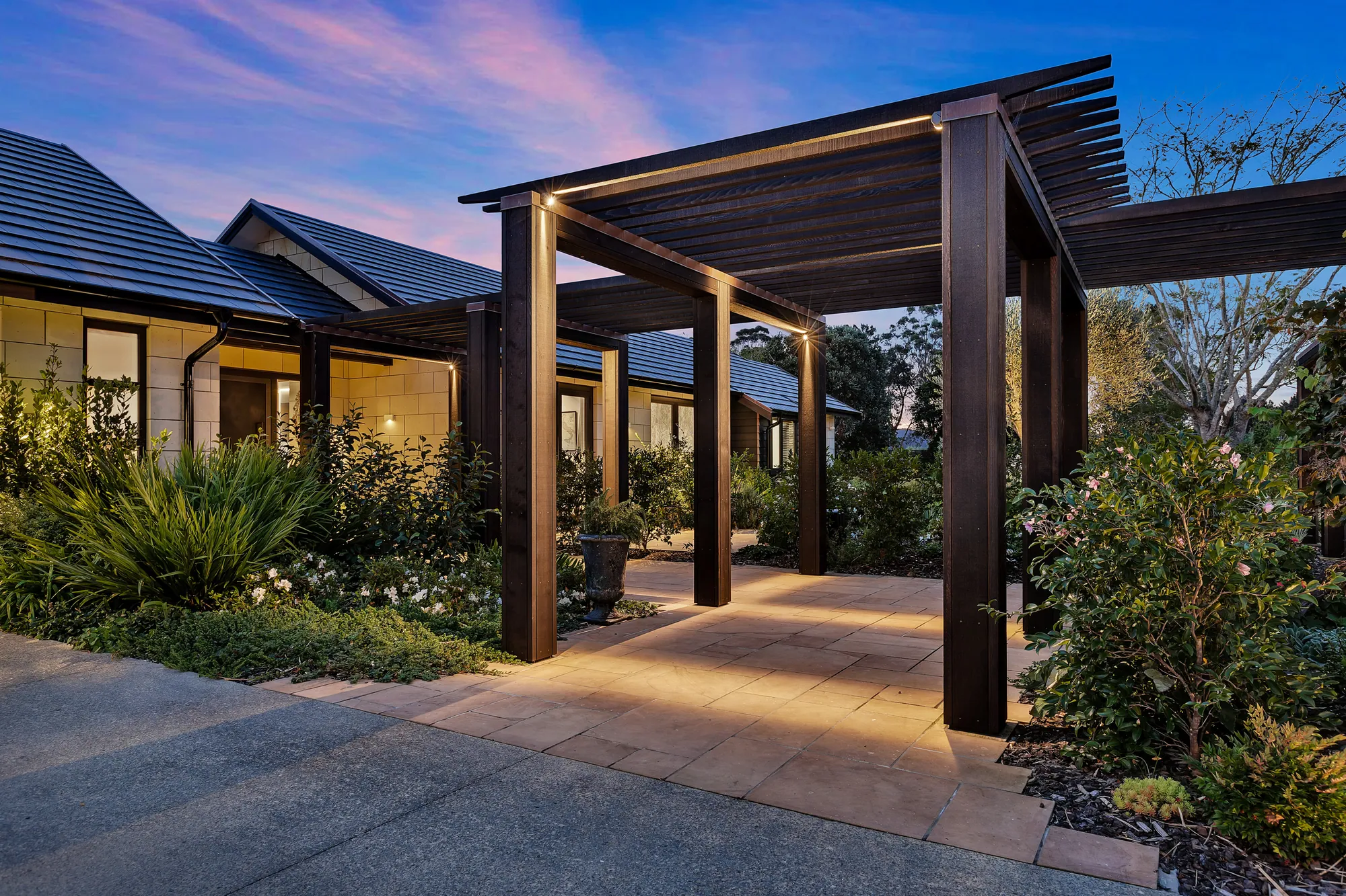 Timber pergola lit at dusk over the path to a stone-clad home
