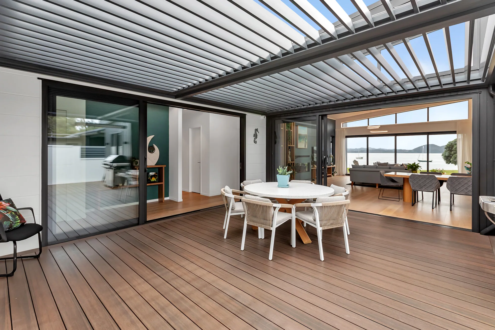 Outdoor dining under a louvred roof, the living room and harbour beyond